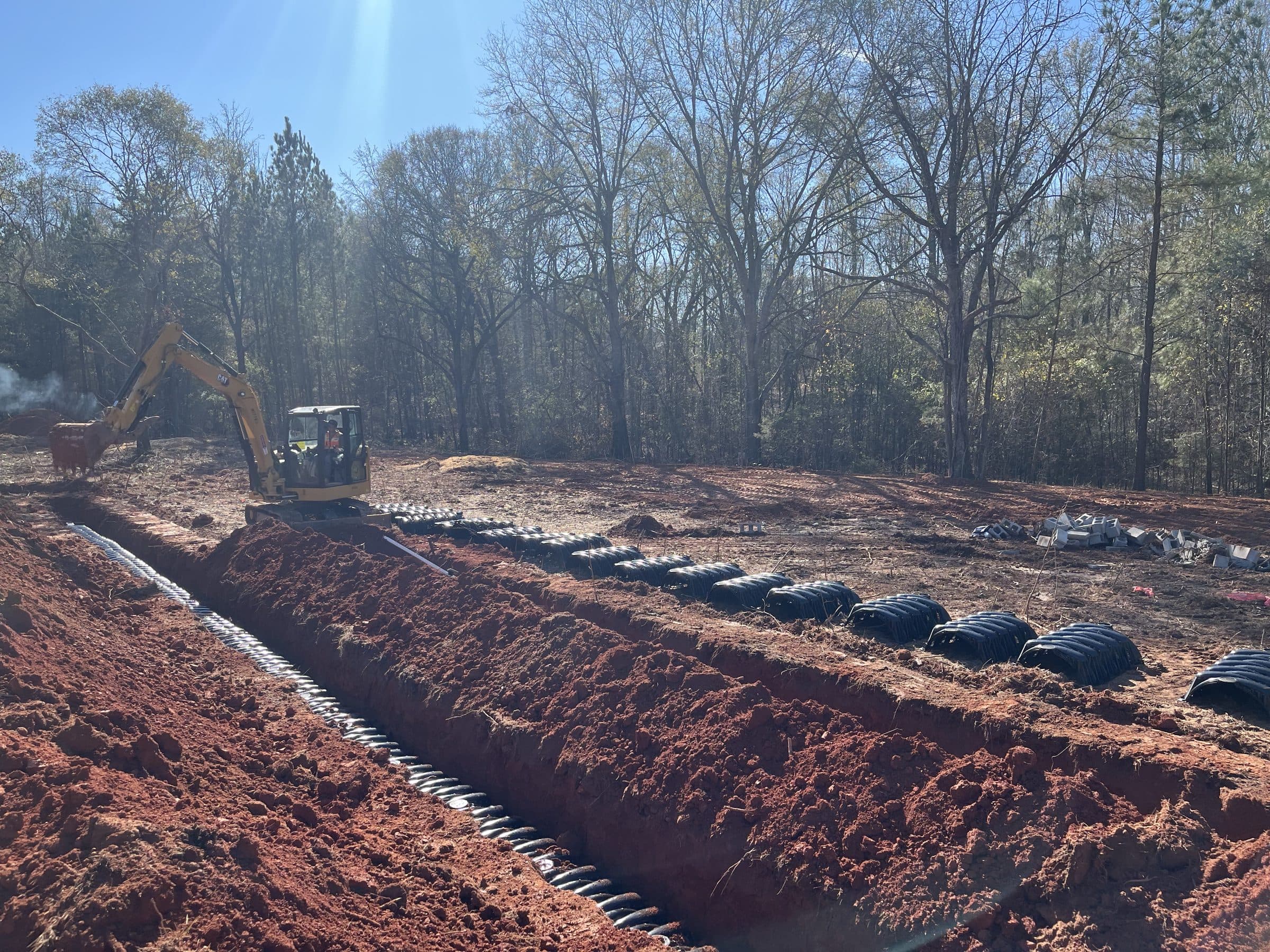 Mini excavator digging drain field trenches with corrugated septic pipe laid in rows of red clay soil